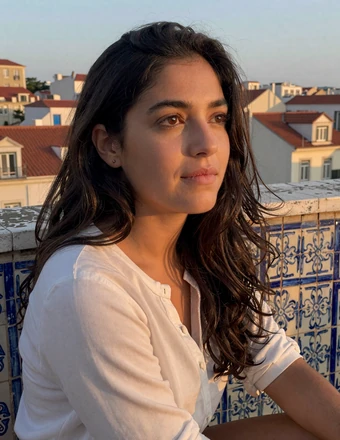 Thoughtful Portuguese woman sitting on a tiled balcony overlooking Lisbon rooftops at sunset with contemplative expression