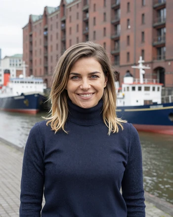 Lena, 28, German woman from Hamburg smiling at the harbor with red brick warehouses behind