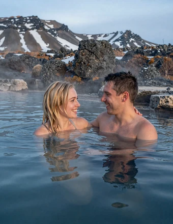 Happy American-Icelandic couple relaxing in a natural geothermal hot spring with snowy Icelandic mountains behind